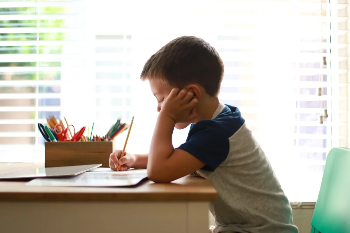 Young boy resting his head on his hand while writing at a desk, looking focused but tired