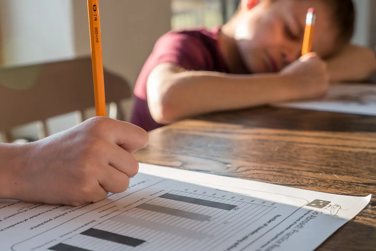 Child resting head on desk while adult supports them with schoolwork, showing overwhelm in learning context